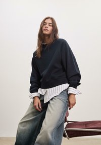 Felpa corta blu marina sopra una camicia bianca a righe. Jeans blu chiaro, larghi e sbiaditi. Design semplice con tessuto morbido e vestibilità rilassata.