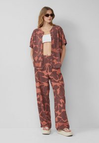 Twee-delige outfit bestaande uit een overhemd met knopen en brede broek in een bruin en verbrand oranjerood abstract patroon, gemaakt van lichte stof.