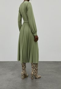 Lång olivgrön klänning med lång ärm, plisserad design och skärp i midjan, kombinerad med knähöga stövlar med ormskinnsmönster.