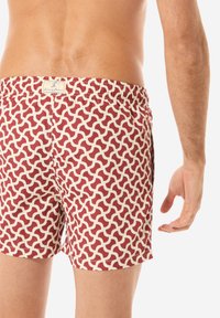Pantaloni da bagno rossi e crema con un motivo ondulato, vita elastica e tasche laterali. Il materiale sembra leggero con una texture liscia.