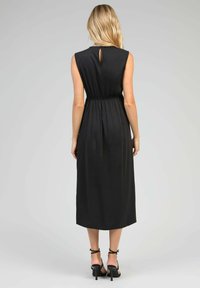 Robe maxi noire sans manches avec détail en trou de clef à l'avant, ceinture élastique et design de jupe fluide. Portée avec des talons noirs à lanières.