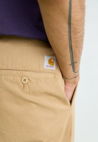 Tan shorts with a smooth texture, featuring a back pocket with a flat button and a square white label with an orange logo.