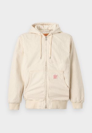 Μπεζ ζακέτα με κουκούλα και φερμουάρ, λεπτές κάθετες μαύρες ρίγες, μπροστινές τσέπες, κορδόνια, ελαστικές μανσέτες και ένα κόκκινο ορθογώνιο ταμπελάκι στην τσέπη.