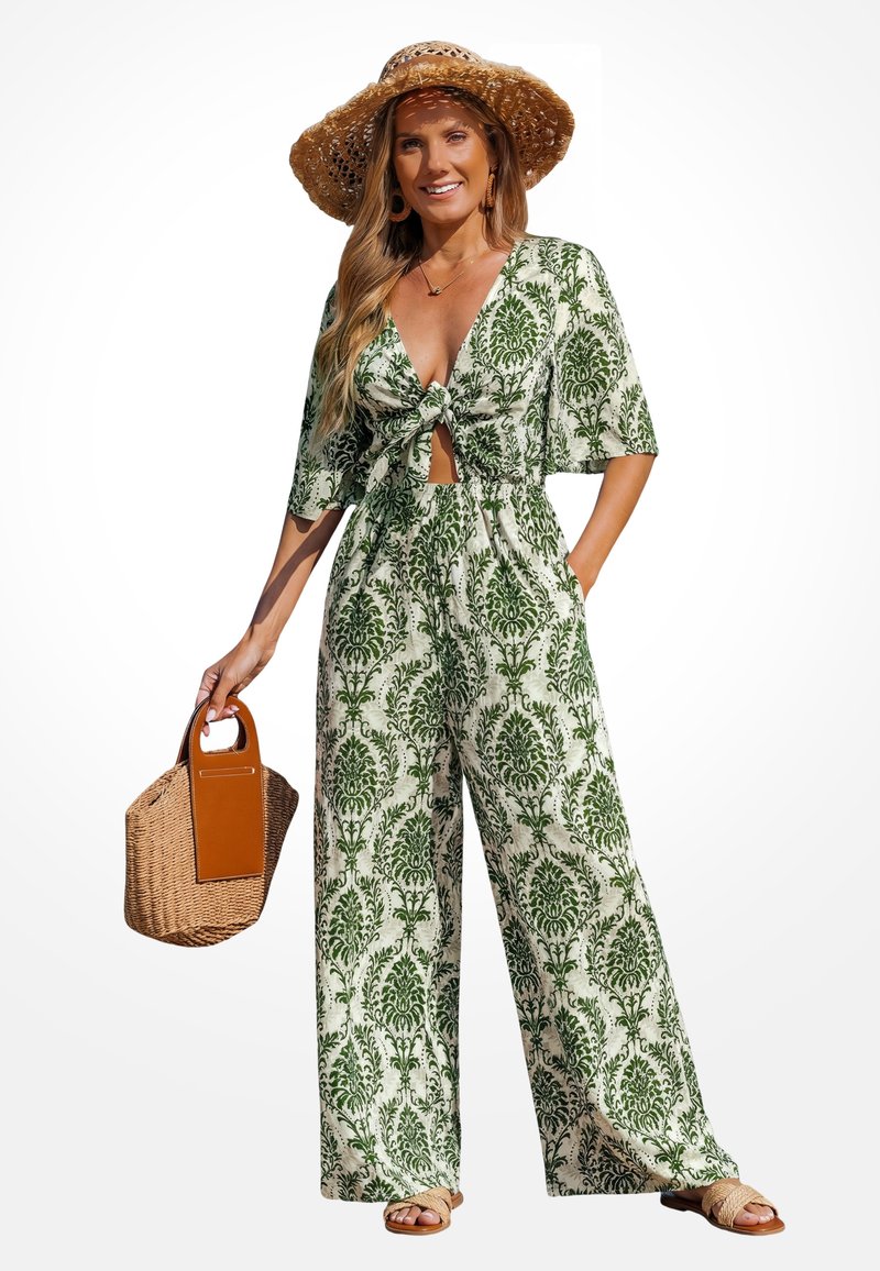 Vrouw in een groen-witte jumpsuit met patroon, met een knoop aan de voorkant, broek met wijd uitlopende pijpen, strooien zonnenhoed, gevlochten handtas, sandalen en lang golvend blond haar.