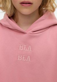 Růžová mikina vyrobená z měkkého materiálu s přední kapsou ve stylu "kangaroo". Má vystouplou výšivku ve světle růžové barvě s nápisem "BLA BLA."