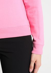 Felpa rosa con orlo e polsini a coste, caratterizzata da una texture morbida. Indossata sopra pantaloni neri aderenti, che mettono in evidenza un design casual.