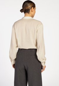 Woman with tied-back hair wearing a loose cream long-sleeve blouse tucked into high-waisted dark gray trousers, facing away.