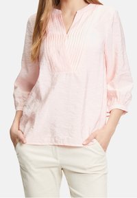 Blouse rose clair à manches longues avec col décolleté et plis verticaux. Le tissu semble doux avec une texture légèrement froissée.