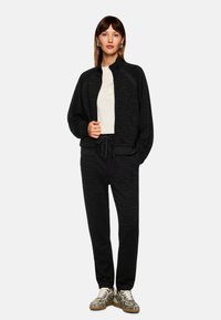 Black zip-up jacket with subtle zebra pattern, paired with matching joggers. Model wears a light top and patterned sneakers.