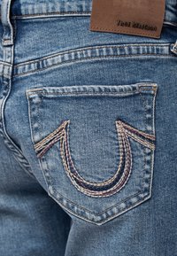 Niebieskie dżinsy z wyraźnym wzorem kieszeni, z wielokolorowym przeszyciem w kształcie litery U. Przypięta brązowa skórzana metka z napisem "TRUE RELIGION".