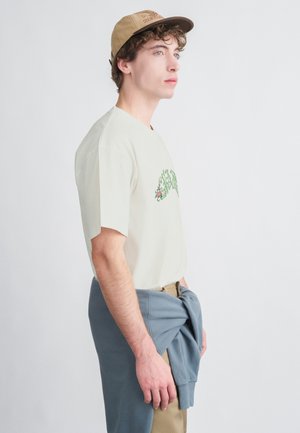 Ανοιχτό μπεζ βαμβακερό t-shirt με πράσινη κεντητή λουλουδάτη διακόσμηση, συνδυασμένο με μπλε φούτερ δεμένο γύρω από τη μέση και μπεζ παντελόνι. Καπέλο στο κεφάλι.