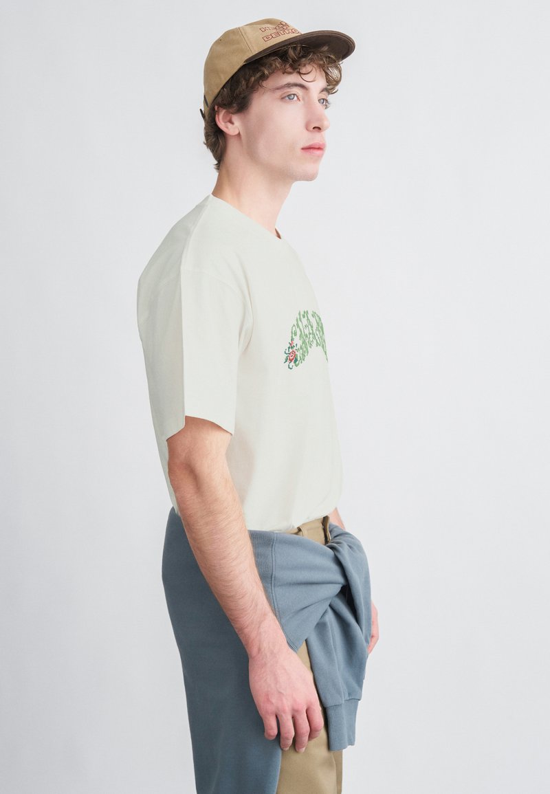 Ανοιχτό μπεζ βαμβακερό t-shirt με πράσινη κεντητή λουλουδάτη διακόσμηση, συνδυασμένο με μπλε φούτερ δεμένο γύρω από τη μέση και μπεζ παντελόνι. Καπέλο στο κεφάλι.