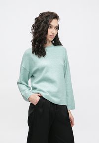 Mintgrønn genser i myk strikk, med rund hals og avslappet passform. Lange ermer med ribbestrikkede mansjetter, kombinert med svarte bukser.