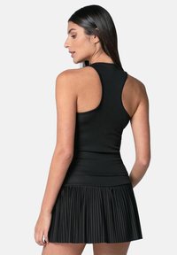 Vrouw met lang donker haar in een zwarte mouwloze racerback-top en een geplooide zwarte mini-rok, die zijwaarts poseert tegen een effen achtergrond.