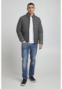 Pilkas paminkštintas švarkas su aukšta apykakle, priekiniais užtrauktukais ir elastingomis rankovėmis, dėvimas ant baltos marškinėlių ir mėlynų džinsų, derinamas su juodais sportbačiais.