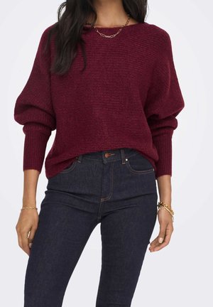 Femme portant un pull en maille texturée bordeaux à manches longues et un jean taille haute bleu foncé, accessoirisée avec des bracelets et un collier en or.