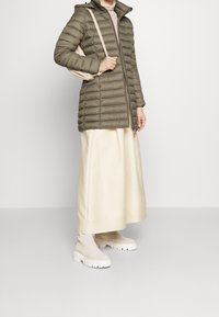 Doudoune verte olive, au design matelassé, avec fermeture éclair et capuche. Associée à une jupe maxi crème et des bottines beiges.