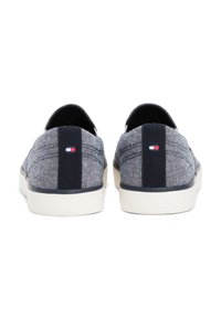 Ett par blå tyg slip-on sneakers med vita sulor och en liten röd, vit och blå logotyp på baksidan av remmen.