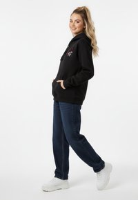 Felpa nera con tasca frontale e grafica rossa, abbinata a jeans blu scuri e sneakers bianche. Il modello è in profilo su uno sfondo bianco.