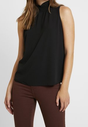 Frau trägt ärmelloses schwarzes Oberteil mit hohem Kragen und braune Hose, steht vor einfachem hellem Hintergrund.