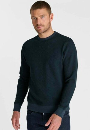 Man with short hair and beard wearing a dark textured long-sleeve sweater and dark pants, standing against a light background.
