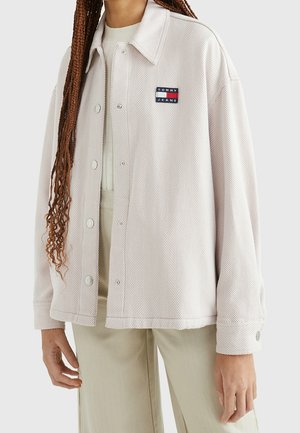 Person iført en cremefarvet fiskebensmønstret jakke med knapper og et Tommy Jeans-logo på brystet samt beige bukser.