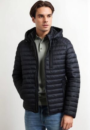 Navy puffer jacket with a hood, featuring quilted texture, zippered pockets, and a front zipper closure. Worn over a light green polo.