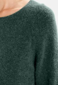 Pull en laine vert foncé avec une surface douce et texturée et un col rond, présentant une coupe ample et des détails de design minimalistes.