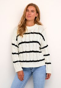 Pulover tricotat cu o bază crem și dungi negre îndrăznețe, având un model texturat și manșete cu dungi. Asortat cu blugi de un albastru deschis.