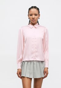 Blusa rosa claro de cetim com gola, mangas compridas e fecho de botões, combinada com saia cinza plissada. Textura suave e corte descontraído.