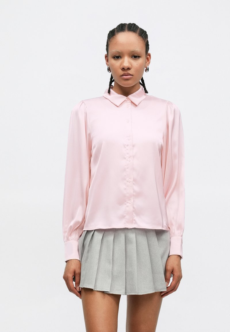 Blusa rosa claro de cetim com gola, mangas compridas e fecho de botões, combinada com saia cinza plissada. Textura suave e corte descontraído.