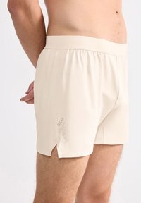Beige bomullskalsonger med elastisk midjeband, sidoflikar och subtil logotyp på nederkanten. Mjuk känsla och åtsittande design.