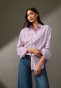 Camicia button-up rosa chiaro con strisce verticali rosse, maniche arrotolate e taschino frontale, abbinata a jeans a gamba larga blu.