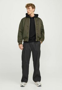 Olive green bomber jacket with zipper, black hooded sweatshirt underneath, paired with black cargo pants and silver athletic shoes.