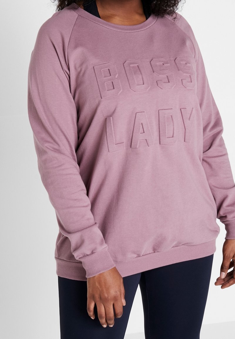 Femme portant un sweat-shirt mauve avec le texte en relief "BOSS LADY" et des leggings bleu marine, debout devant un fond clair uni.