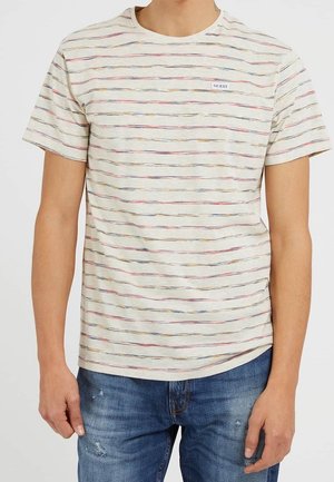 Mężczyzna ubrany w kremowy t-shirt z krótkim rękawem z cienkimi, wielokolorowymi poziomymi paskami i małym logo Guess, w połączeniu z niebieskimi, przetartymi jeansami.