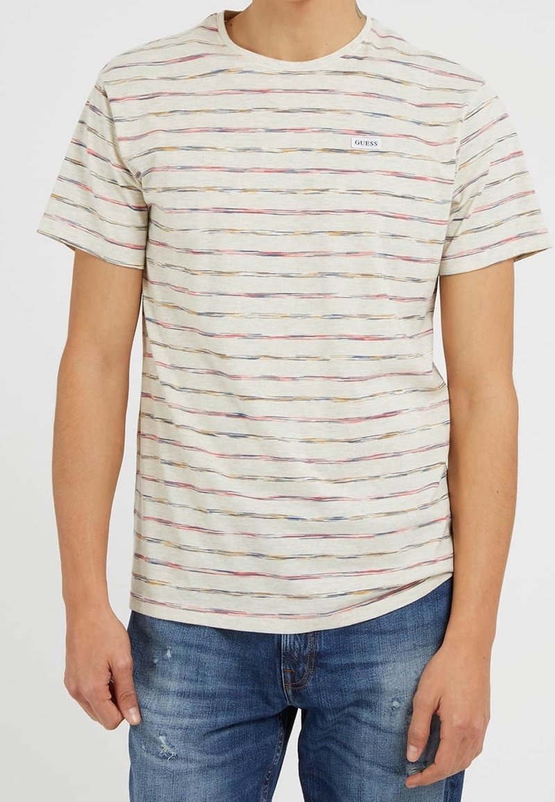 Homme portant un T-shirt crème à manches courtes avec de fines rayures horizontales multicolores et un petit logo Guess, associé à un jean bleu déchiré.