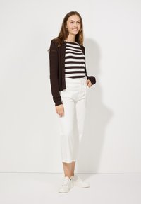 Braune, Strickjacke über einem schwarz-weißen gestreiften Oberteil, kombiniert mit weißen, knielangen Hosen und weißen Turnschuhen, vor einem einfach gehaltenen Hintergrund.