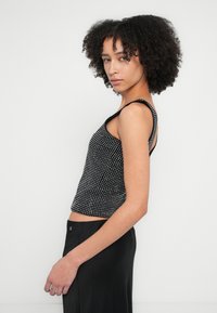 Črna cepljena majica brez rokavov z belimi pikami in oprijetim krojem. Ima tanke naramnice in je kombinirana s črno krilo, prikazano z bočnega vidika.