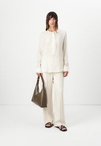 Lys kremfarget bluse med knytehals og teksturerte detaljer, sammen med off-white vide bukser. Holder en mørk olivengrønn håndveske. Svarte sandaler.