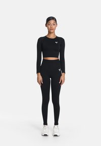 Čierny dlhý rukáv crop top s logom, spárovaný s čiernymi legínami s vysokým pásom. Materiál vyzerá hladko a pružne; prítomné biele akcenty.