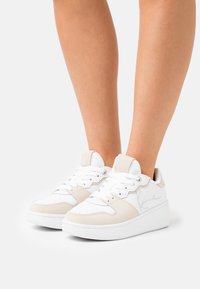 Hvite og beige sneakers med overdel i glatt lær, perforert tå og polstret såle. Har snøring og subtil merkevare.