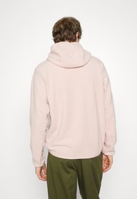 Felpa con cappuccio in pile rosa con ampio cappuccio, maniche lunghe e vestibilità comoda. Presenta polsini a costine e una texture liscia. Indossata con pantaloni verdi.