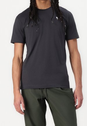 Mand iført en sort t-shirt med korte ærmer med et lille hvidt fuglelogo og mørkegrønne bukser med snøre foran mod en hvid baggrund.