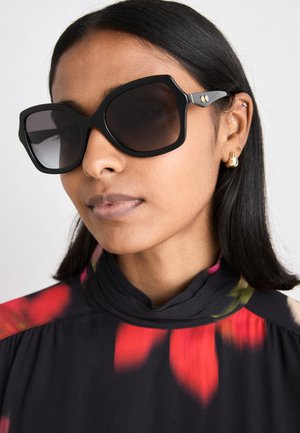 Czarne, duże okulary przeciwsłoneczne o geometrycznej oprawie, ciemne soczewki i złote akcenty, noszone z topem w kwiatowy wzór i wysokim kołnierzem.