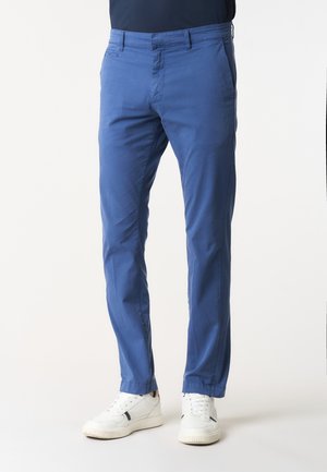 Uomo che indossa pantaloni blu slim fit e sneakers bianche, in piedi davanti a uno sfondo chiaro e semplice.