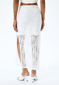 Jupe maxi blanche en dentelle avec des fentes sur les côtés, présentant des motifs floraux complexes et une texture douce. Portée avec des mules à talons blancs.