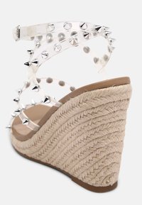 Sandale compensée avec talon en jute tressée et brides transparentes ornées de pointes argentées entourant la cheville.