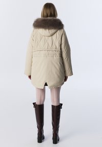 Beige gewatteerde jas met een capuchon en een bruine bontkraag, voorzien van een diamantvormig quiltpatroon en een hoge-lage zoom. Ggedragen met een patroonlegging en kniehoge laarzen.