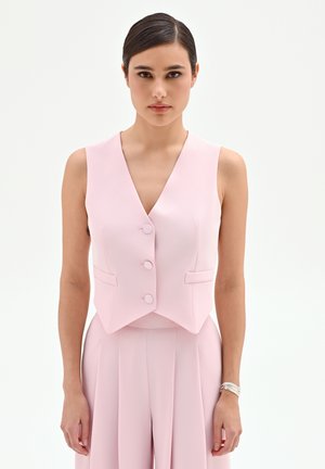 Donna che indossa un gilet smanicato rosa chiaro con bottoni e pantaloni coordinati a vita alta, in piedi davanti a uno sfondo bianco uniforme.